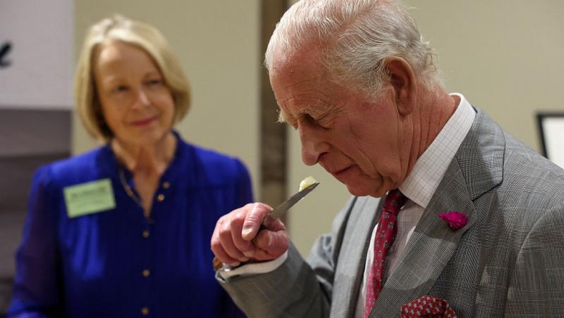 Regele Charles îmbrăcat cu un costum gri, mirosind o bucată de brânză