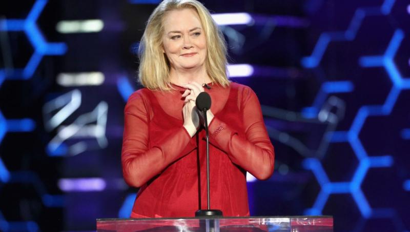 Cybill Shepherd îmbrăcată într-o rochie roșie, vorbind la microfon