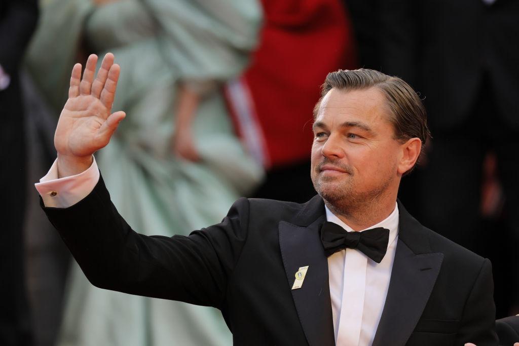Leonardo DiCaprio îmbrăcat într-un costum negru și o cămașă albă, salutând lumea la un eveniment