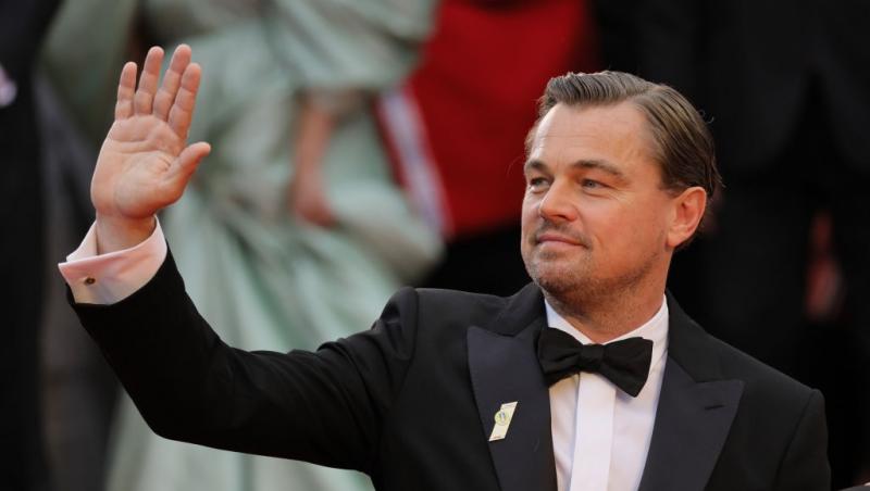 Leonardo DiCaprio îmbrăcat într-un costum negru și o cămașă albă, salutând lumea la un eveniment
