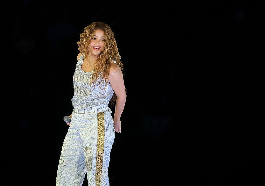 Shakira pe scenă, îmbrăcată cu un maiou alb strălucitor cu aurii și pantaloni asortați