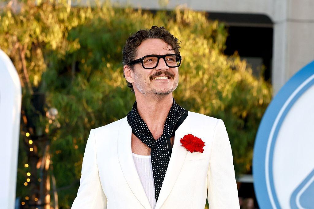 Pedro Pascal îmbrăcat într-un costum alb, accesorizat cu o eșarfă neagră și o floare roșie