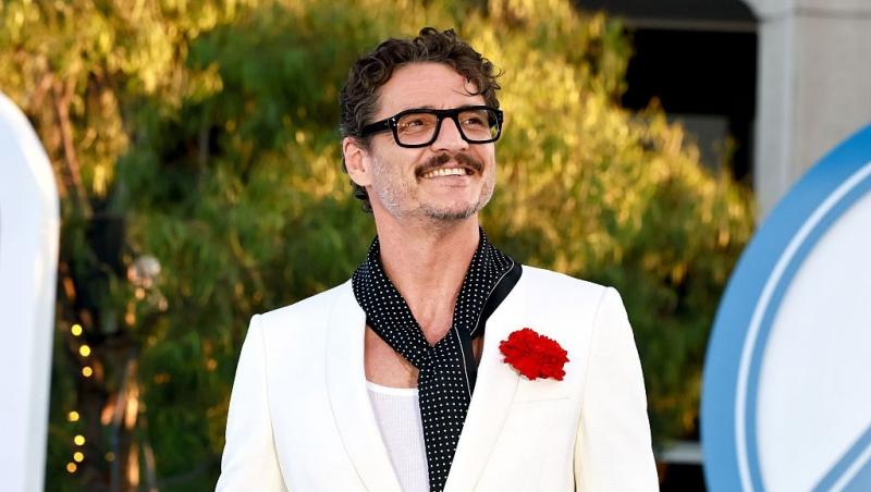 Pedro Pascal îmbrăcat într-un costum alb, accesorizat cu o eșarfă neagră și o floare roșie