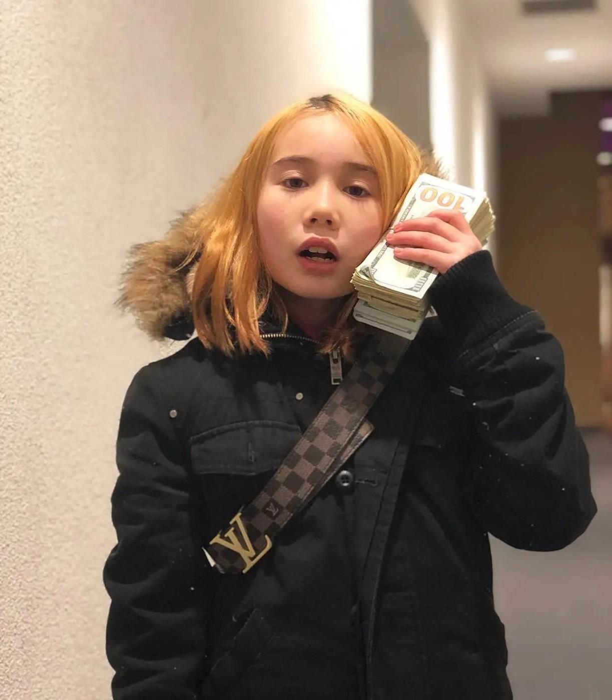 Lil Tay îmbrăcată cu o geacă neagră ținând un teanc de bani la ureche