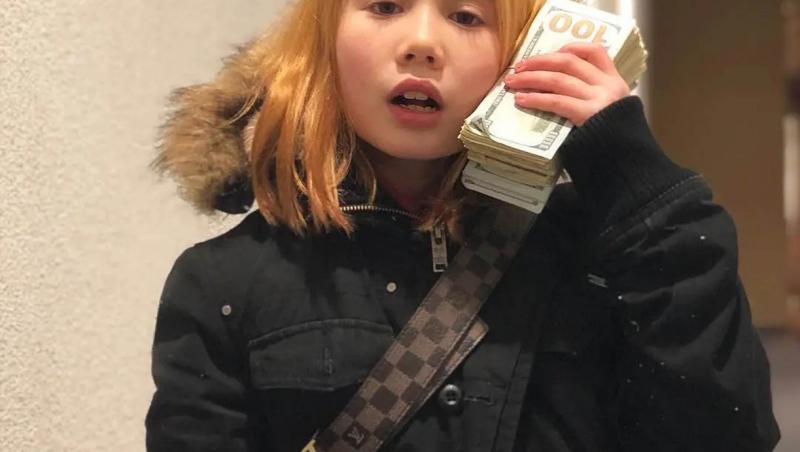 Lil Tay îmbrăcată cu o geacă neagră ținând un teanc de bani la ureche