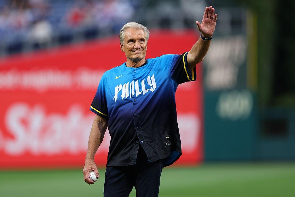 Dolph Lundgren îmbrăcat într-un tricou negru, pe un teren de sport, salutând mulțimea