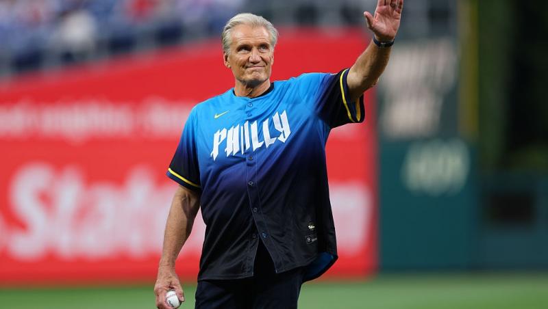 Dolph Lundgren îmbrăcat într-un tricou negru, pe un teren de sport, salutând mulțimea