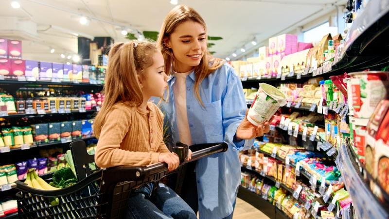 mamă care &icirc;i citește ingredintele de pe un produs copilei sale &icirc;n supermarket
