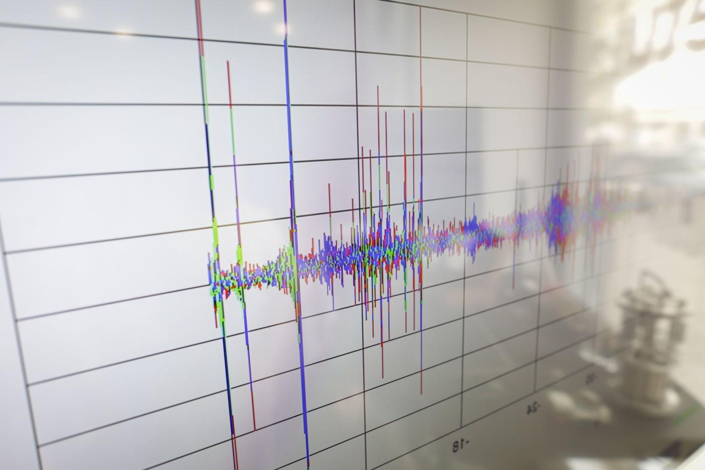 Detalii cu profunzime mică a câmpului (focalizare selectivă), surprinzând oscilații pe o seismogramă afișată pe un ecran digital, cu reflexia unui dispozitiv vizibilă în partea dreaptă.