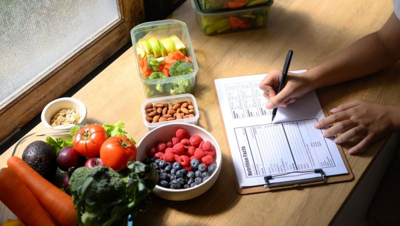femeie care scrie &icirc;ntr-un carnet pe un blat l&acirc;ngă legume și fructe