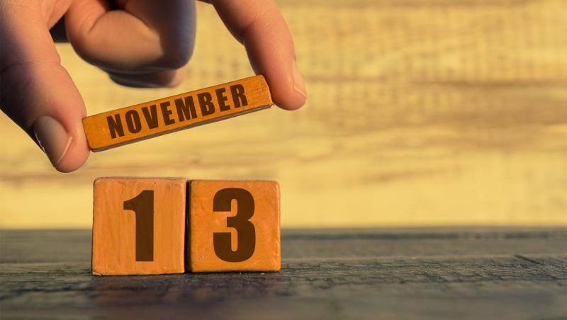 Ziua 13 a lunii, calendar pe un cub de lemn. O m&acirc;nă de femeie așază numele lunii &icirc;n dreptul numărului zilei, pe un fundal din lemn.